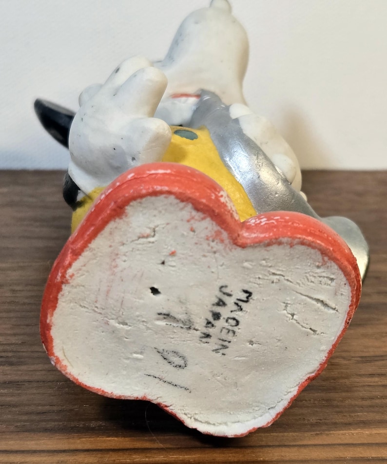 May include: A vintage figurine of a cartoon character, featuring a white body, black ears, and a yellow and gray outfit. The base is red and the bottom reads "MADE IN JAPAN".
