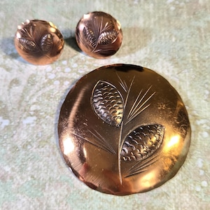 May include: A vintage copper jewelry set featuring a large round brooch and two matching clip-on earrings. Each piece has an embossed design of stylized pinecones and foliage. The set has a warm, metallic finish.