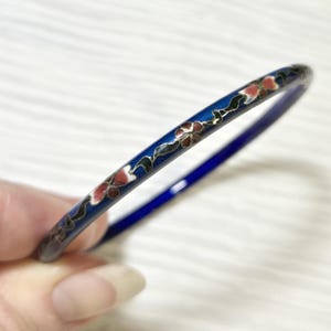Puede incluir: Una pulsera de brazalete de esmalte azul con un diseño floral en rojo, blanco y verde. La pulsera tiene un fondo azul oscuro y un acabado brillante. El patrón floral se repite alrededor de la pulsera.