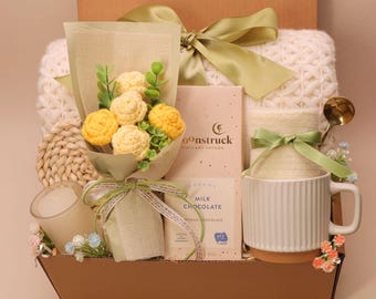 May include: A gift box filled with a white knitted blanket, a bouquet of yellow flowers, a white mug, a candle, and a bar of milk chocolate. The box is tied with a green ribbon.
