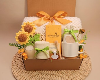 May include: A gift basket containing a white knit blanket with a yellow ribbon, a Moonstruck chocolate bar, a white ribbed mug, a small jar of honey with a wooden dipper, a candle, a fabric pouch, and two crocheted sunflowers with green leaves.