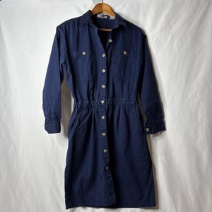 May include: A navy blue long-sleeve dress with a collared neckline and button-down front. The dress features two chest pockets, a cinched waist, and side pockets. It is hanging on a wooden hanger against a white background.