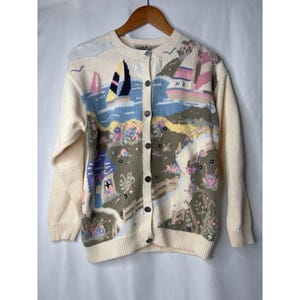 May include: Cream-colored cardigan sweater with a nautical scene design. The button-down sweater features sailboats, houses, and floral details in pastel colors. The long-sleeved sweater is hanging on a wooden hanger.