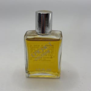 May include: A rectangular glass bottle of Cachet cologne with a silver cap, filled with a golden-yellow liquid. The label on the back is visible through the clear glass, displaying the text Cachet Cologne and 1/2 FL OZ in clear lettering.