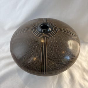 Puede incluir: Un recipiente de cerámica negra, redondo y de perfil bajo, con una pequeña abertura central. La superficie está decorada con intrincados patrones geométricos de líneas finas en color tostado claro, con líneas radiantes y formas triangulares.