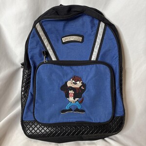 Może przedstawiać: Niebieski plecak z czarnymi detalami i haftem postaci Taza na przedniej kieszeni. Srebrne paski odblaskowe z przodu. Mała prostokątna naszywka znajduje się nad przednią kieszenią. Plecak posiada komorę główną zapinaną na zamek i boczne kieszenie.