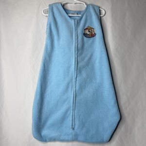 May include: A light blue baby sleep sack with a full-length zipper and a small embroidered patch featuring cartoon characters. The sleep sack is made of a soft, fleece-like material and hangs on a white hanger.