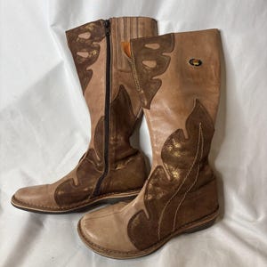 May include: A pair of knee-high boots featuring a combination of light tan leather and dark brown metallic suede overlays. The boots have a side zipper closure, a rounded toe, and a small oval logo patch near the top collar with visible stitching.