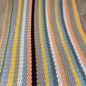 Pode incluir: Um cobertor de crochê feito à mão com listras verticais em várias cores pastel, incluindo pêssego, amarelo, lavanda e cinza. Uma faixa roxa escura percorre o centro. O cobertor é feito com um ponto texturizado.