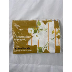 May include: A packaged double flat sheet by Tastemaker. The sheet features a white background with a floral pattern of daisies and orange vertical stripes. The packaging is mustard yellow with text.