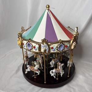 May include: A decorative carousel with a striped canopy in red, white, purple, green, and gold. The carousel features several small animal figurines, including horses and giraffes, and is set on a dark brown base.