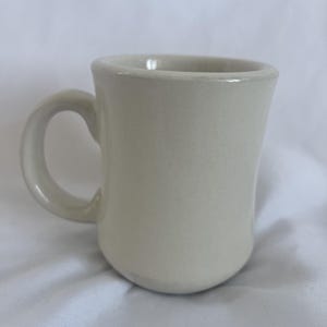 May include: A cream-colored ceramic mug with a rounded body and a curved handle. The mug has a smooth, glossy finish and appears to be empty. The mug is sitting on a white surface.