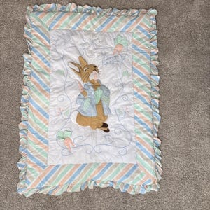 May include: A rectangular baby quilt featuring Peter Rabbit. The center has an embroidered image of Peter Rabbit holding a carrot. The quilt has a white background with a striped border in pastel colors and a ruffled edge.