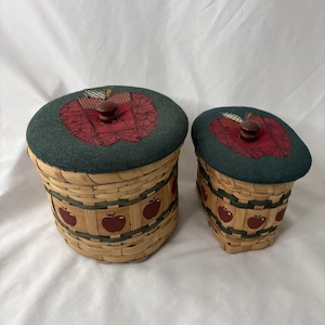 May include: Two woven wicker baskets with lids. The lids are dark green with a red apple design. The baskets have a repeating apple pattern around the sides. The lids have a wooden knob. The baskets are different sizes.