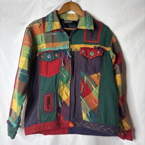 Puede incluir: Chaqueta patchwork multicolor con paneles rojos, verdes, amarillos y azules. La chaqueta tiene cremallera, varios bolsillos y costuras decorativas. Colgada en una percha de madera.