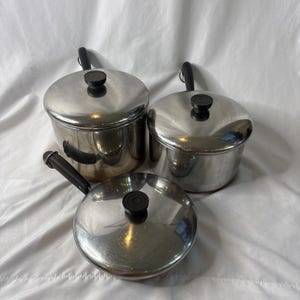 May include: A set of three stainless steel cooking pots with matching lids. Each pot features a polished silver finish, a black handle, and a black knob on the lid. The set includes one deep pot with a side handle and two smaller pots with long handles.