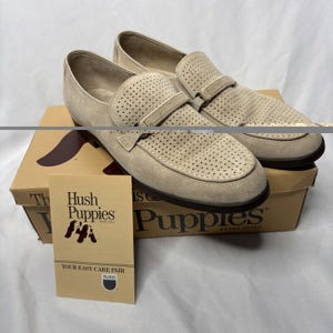 May include: A pair of tan Hush Puppies loafers with perforated detailing on the vamp and a strap across the top. The shoes sit on a brown shoe box with the Hush Puppies logo and a small card that says "YOUR EASY CARE PAIR".