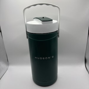 May include: A dark green insulated water jug with a white handle and lid. The jug has the text "HUDSON'S" printed on the front. The lid has a small, dark green flip-top opening. The handle is white and arched.