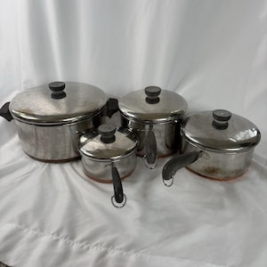 Puede incluir: Un juego de cuatro ollas de acero inoxidable con tapas y asas negras. Las ollas varían en tamaño, con una base de color cobre. Las tapas tienen perillas negras. Sobre una superficie blanca.