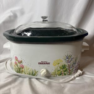May include: A white Rival Crock-Pot stoneware slow cooker with a dark green rim and a clear glass lid. The exterior features a botanical print of clover and dill. It has a single front-facing control knob for Off, Low, and High settings.