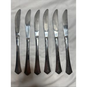 Könnte beinhalten: Sechs Tafelmesser aus Edelstahl mit gebogenen Klingen. Die Messer haben eine polierte Oberfläche und ein dekoratives Griffdesign. Die Klingen sind etwa 23 cm lang, und die Griffe haben eine ausgestellte, verzierte Basis.