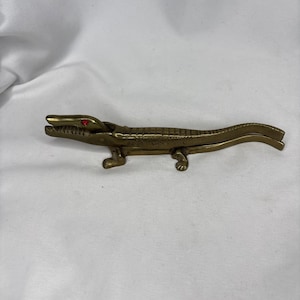 May include: A brass alligator-shaped nutcracker with red eyes. The nutcracker is detailed with scales and has a hinged jaw. The alligator is lying down on a white surface.