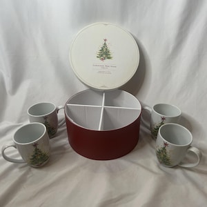 Puede incluir: Un juego de cuatro tazas de Navidad de cerámica blanca con un diseño de árbol de Navidad, una caja dividida roja y blanca y una tapa redonda con una ilustración de árbol de Navidad.