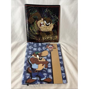 May include: Two Tazmanian Devil themed notebooks. One notebook has a black cover with a colorful Taz graphic. The other notebook has a blue denim-like cover with a Taz graphic and a wooden-like accent.