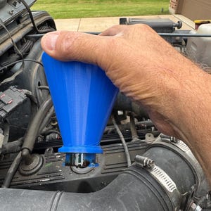 May include: A bright blue funnel is held over an engine component. The funnel is made of plastic and has a wide mouth that tapers down to a narrow spout. The engine is partially visible, with various hoses and components surrounding the funnel.