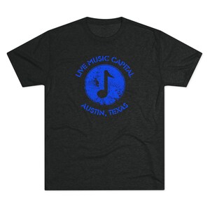 May include: Dark gray t-shirt with a distressed blue graphic. The graphic features a musical note inside a circle, with the words "LIVE MUSIC CAPITAL" above and "AUSTIN, TEXAS" below. A casual tee for music lovers.
