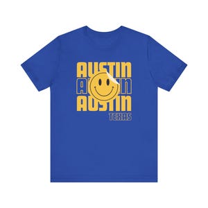 May include: Royal blue t-shirt featuring a yellow smiley face and the word "AUSTIN" stacked three times. Below the smiley face is the word "TEXAS" in a smaller font. The shirt is a short-sleeved crew neck.
