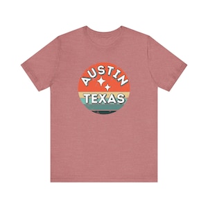 May include: A short-sleeved, mauve t-shirt featuring a circular graphic with the words "AUSTIN TEXAS" in white, set against a retro-style design of orange, yellow, teal, and black stripes. The shirt is made of a soft material.