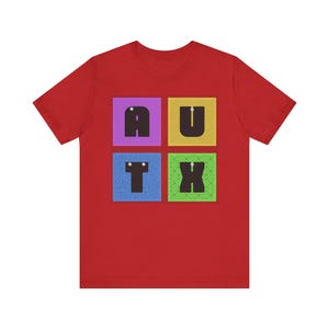 May include: Red t-shirt with the letters A, U, T, and X arranged in a square pattern. Each letter is in a different colored square: purple, yellow, blue, and green. The shirt is made of a soft material.