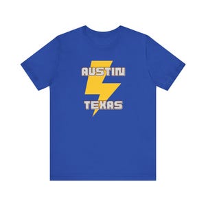 May include: Royal blue t-shirt featuring a yellow lightning bolt graphic with the words "AUSTIN TEXAS" in a white and yellow font. The shirt is a short-sleeved crew neck.