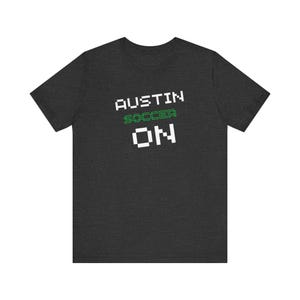 May include: Dark gray t-shirt with the words "AUSTIN" in white, "SOCCER" in green, and "ON" in white. The text has a pixelated, retro video game style. A casual tee for soccer fans.