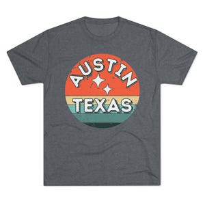 May include: Dark gray t-shirt featuring a retro design with the text "AUSTIN TEXAS" in a circular graphic. The graphic includes orange, yellow, teal, and white colors, with two star shapes.