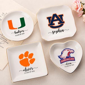 May include: A collection of personalized ceramic ring dishes. The dishes are white with gold trim and feature various designs, including college logos and names. The text includes names like "Hudson", "Sophia", "Oliver", and "Olivia", with "Class of 2026" below each name.