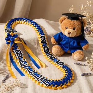 May include: A graduation lei in blue and gold with the name "BELLA" and the year "2025" in black and white lettering. A teddy bear wearing a graduation gown and cap sits nearby. The lei has a "Class of" tag.