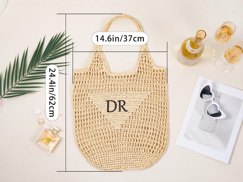 May include: A beige woven tote bag with a heart-shaped design and the initials "DR". The bag is 14.6 inches wide and 24.4 inches tall. The image also includes a bottle of champagne, sunglasses, and perfume.