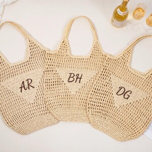 May include: Three beige woven tote bags with open weave construction. Each bag has a handle and a pocket with embroidered initials: "AR", "BH", and "DG". The bags are arranged on a white surface with a bottle of wine and glasses.