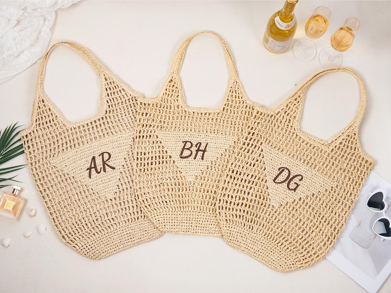 May include: Three beige woven tote bags with open weave construction. Each bag has a handle and a pocket with embroidered initials: "AR", "BH", and "DG". The bags are arranged on a white surface with a bottle of wine and glasses.