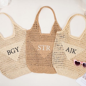 May include: Three woven tote bags in beige and tan hues. Each bag features a unique monogram: "BGY", "STR", and "AJK". The bags have open weave construction and sturdy handles. A pair of pink heart-shaped sunglasses rests on one bag.