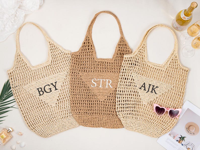 May include: Three woven tote bags in beige and tan hues. Each bag features a unique monogram: "BGY", "STR", and "AJK". The bags have open weave construction and sturdy handles. A pair of pink heart-shaped sunglasses rests on one bag.