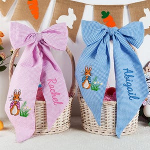 May include: Two Easter baskets with gingham bows. One basket has a pink bow with "Rachel" embroidered, the other has a blue bow with "Abigail" embroidered. Each bow features an embroidered bunny and sits atop a wicker basket.