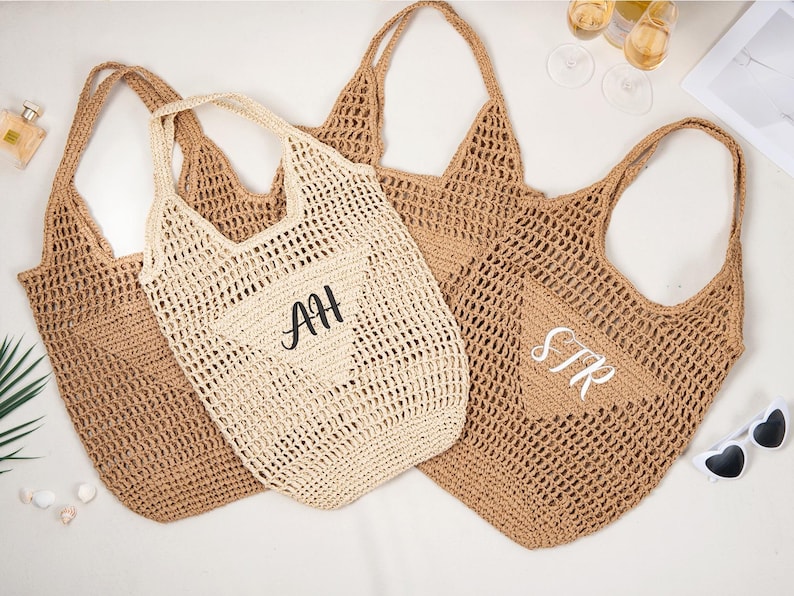 May include: Three woven tote bags in shades of tan and cream. One bag features the initials "AH" in black, and another has "STR" in white. The bags have long handles and a netted design, perfect for beach or market.