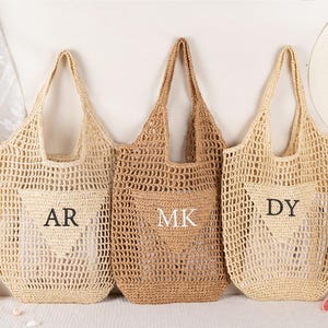 May include: Three crocheted tote bags in varying shades of beige and tan. Each bag features a pocket with embroidered initials: "AR", "MK", and "DY". The bags have long handles and a woven, open-weave design, perfect for a beach or market.