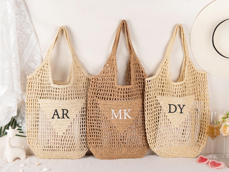 May include: Three crocheted tote bags in varying shades of beige and tan. Each bag features a pocket with embroidered initials: "AR", "MK", and "DY". The bags have long handles and a woven, open-weave design, perfect for a beach or market.