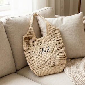 May include: A beige crocheted tote bag with a heart-shaped design and the embroidered letters "BK". The bag has a handle and is resting on a beige sofa with pillows and a blanket.