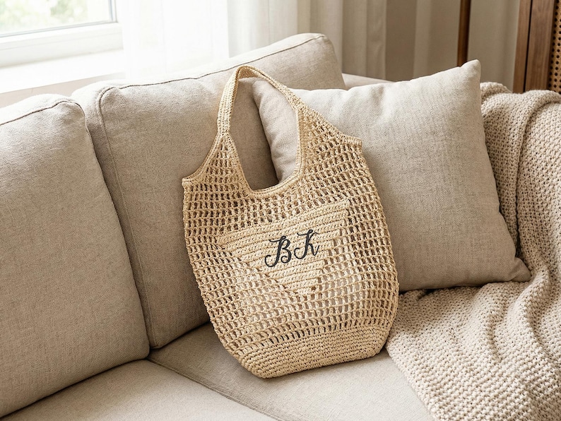 May include: A beige crocheted tote bag with a heart-shaped design and the embroidered letters "BK". The bag has a handle and is resting on a beige sofa with pillows and a blanket.