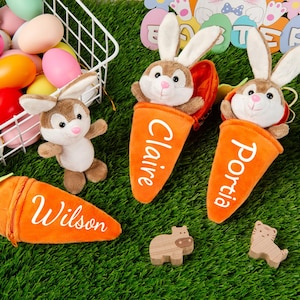 Può includere: Immagine a tema pasquale con coniglietti di peluche, sacchetti a forma di carota con nomi e figure decorative di animali in legno. I coniglietti sono marroni e bianchi e le sacche sono arancioni con scritte bianche.